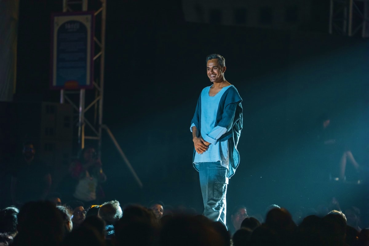 Man in blue shirt performing on stage under bright spotlight with crowd silhouettes in foreground at live event venue