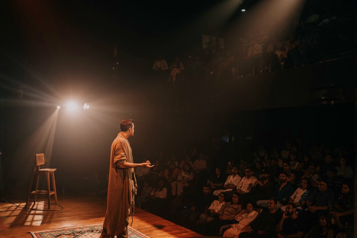 Speaker in orange shawl performing on stage under spotlights with audience seated around wooden theater floor in Dubai