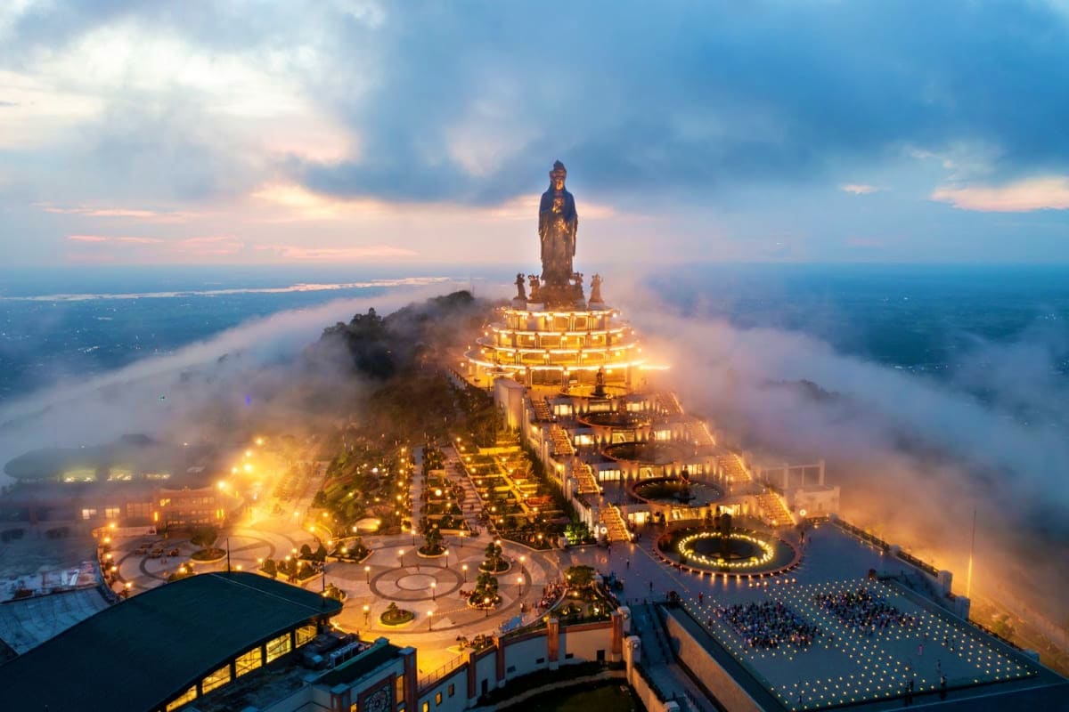 Illuminated statue of Bodhisattva Quan Am at Ba Den Mountain temple complex at dusk, surrounded by misty landscape and golden