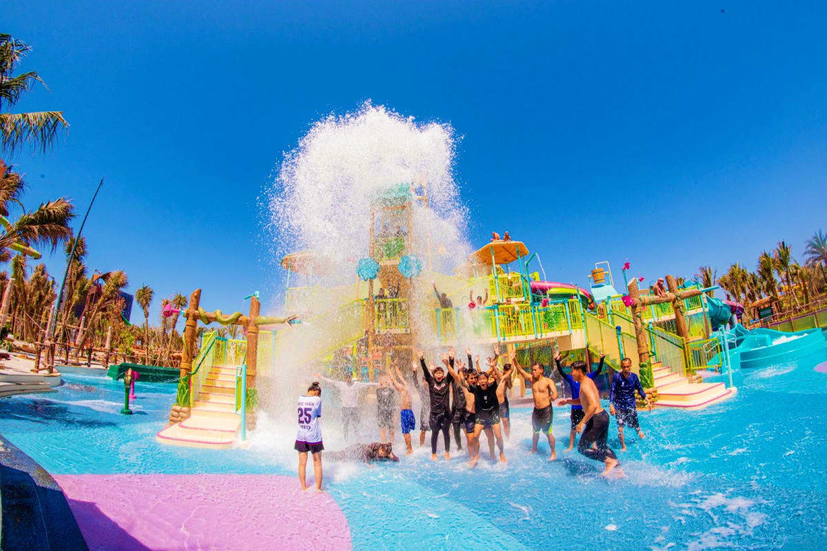Large water bucket tipping over excited visitors at colorful water park playground with palm trees and blue sky at Sun World