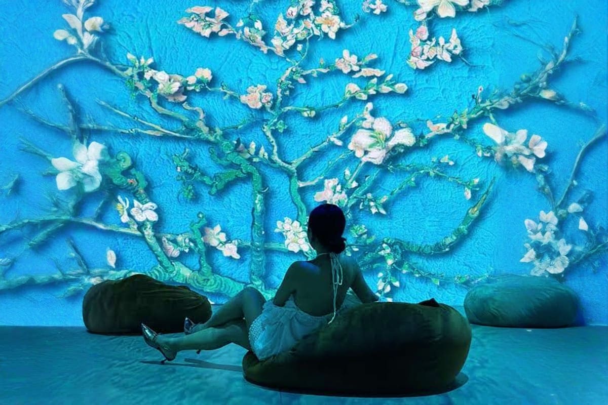 Metashow Exhibition Two visitors sitting on black bean bag chairs viewing a large blue wall mural featuring white almond blossoms and green branc