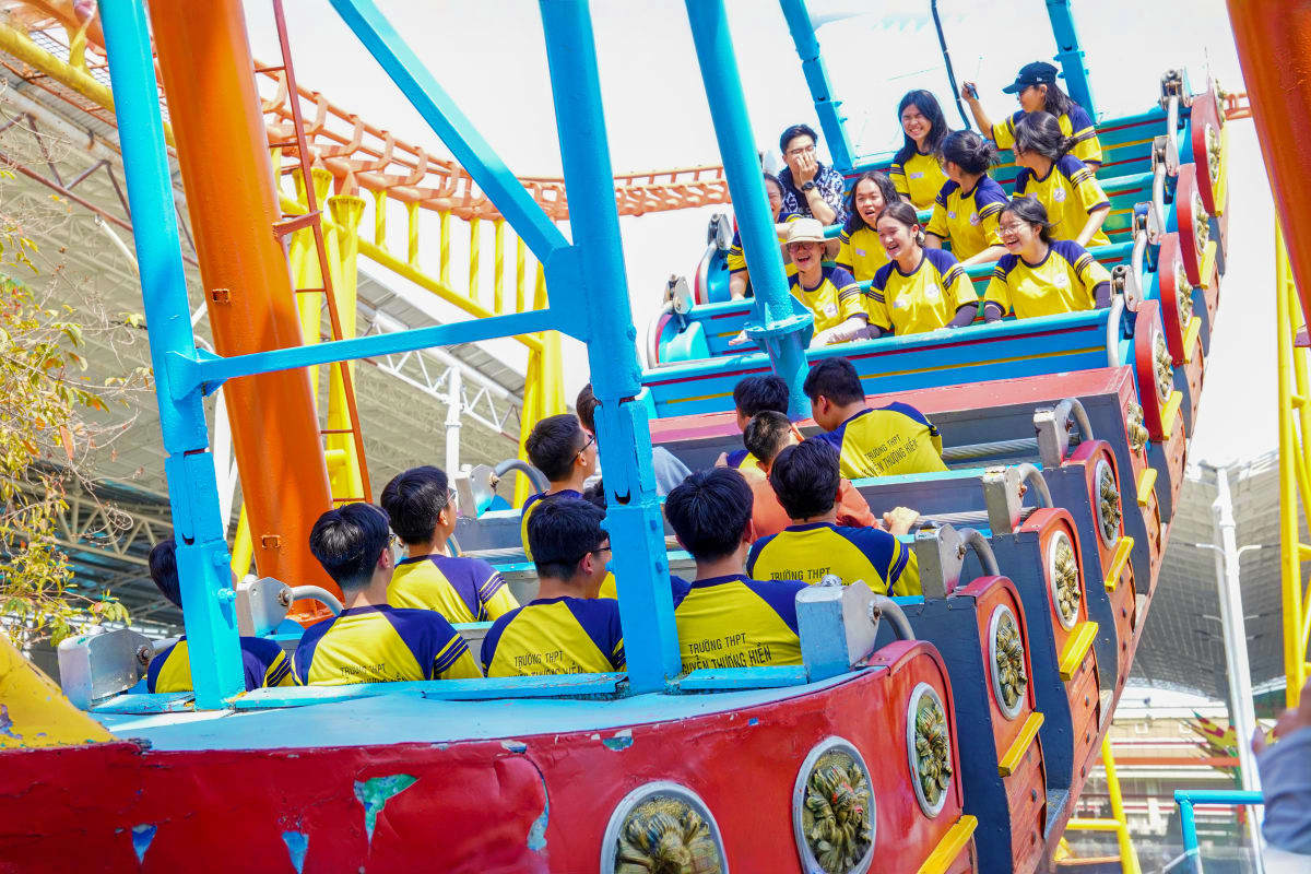 Group of visitors wearing yellow vests riding a colorful roller coaster with blue and orange support structures at Suoi Tien