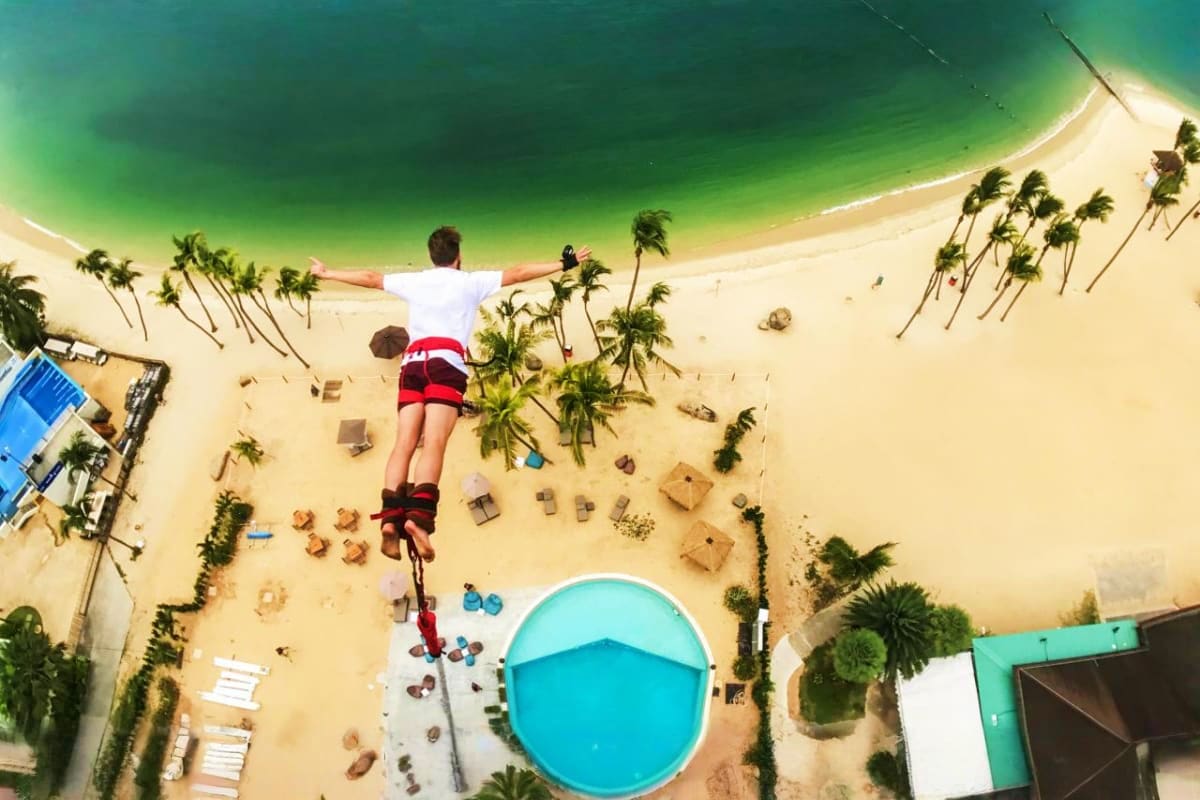 AJ Hackett Sentosa Aerial view of person bungee jumping above tropical beach with palm trees and turquoise water at AJ Hackett Sentosa