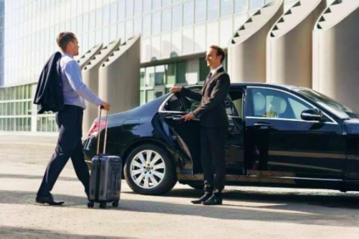 Businessman with luggage greeting driver at black sedan outside modern office building for airport drop-off service