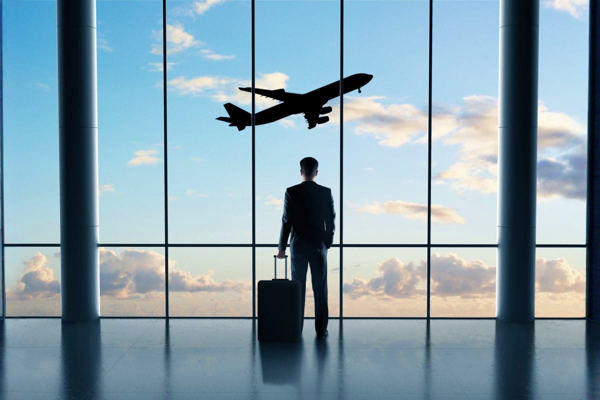 Businessman in suit standing at airport terminal window with luggage, watching airplane take off against blue sky