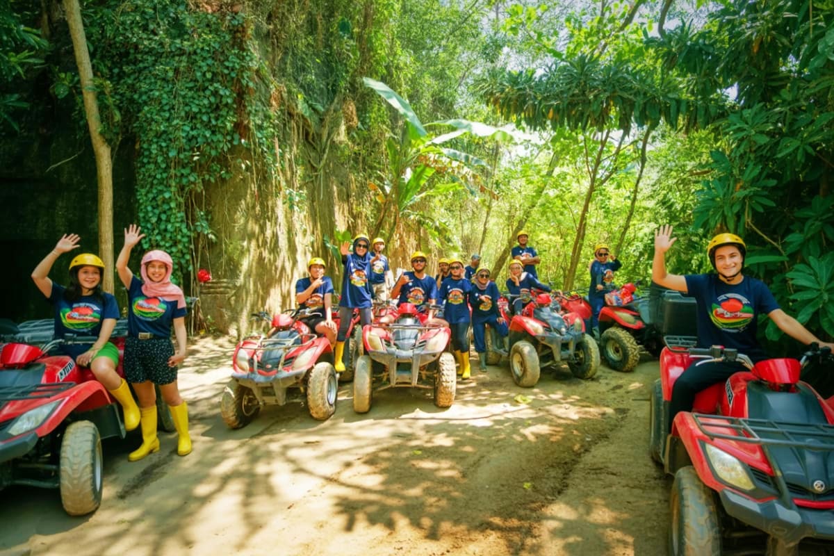 Bali ATV Tour Large group of tourists wearing yellow helmets and matching blue shirts riding red ATVs on a shaded jungle trail in Bali