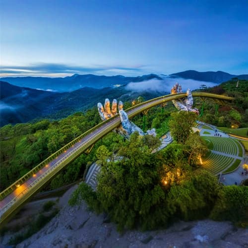 Golden Hand Bridge spanning lush mountains at dusk, Bana Hills landmark with illuminated stone hands and forested valley land