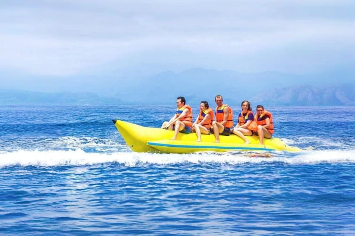 Banana Boat Ride in Dubai 15 Min Group of six people wearing orange life jackets sitting on a bright yellow banana boat riding waves in blue ocean with mounta