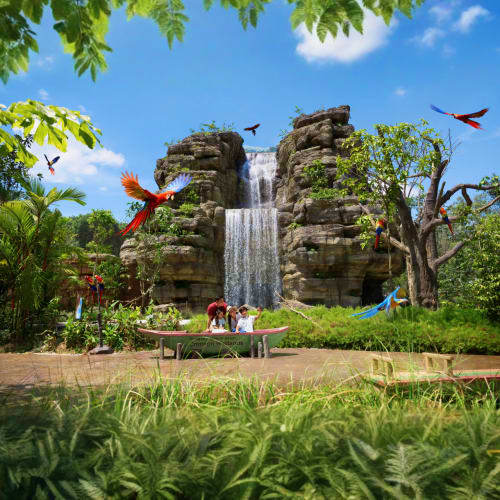 Tropical waterfall attraction with colorful macaws flying, lush greenery and rock formations with visitors viewing the scenic