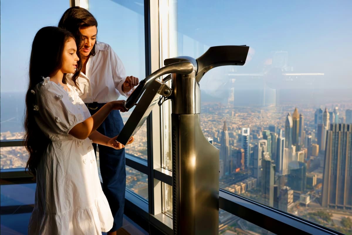 Two visitors using a viewing telescope on an observation deck overlooking the Dubai cityscape from Burj Khalifa