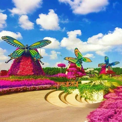 Colorful butterfly sculptures with pink flowers in a lush garden setting under blue sky with white clouds at Butterfly Garden