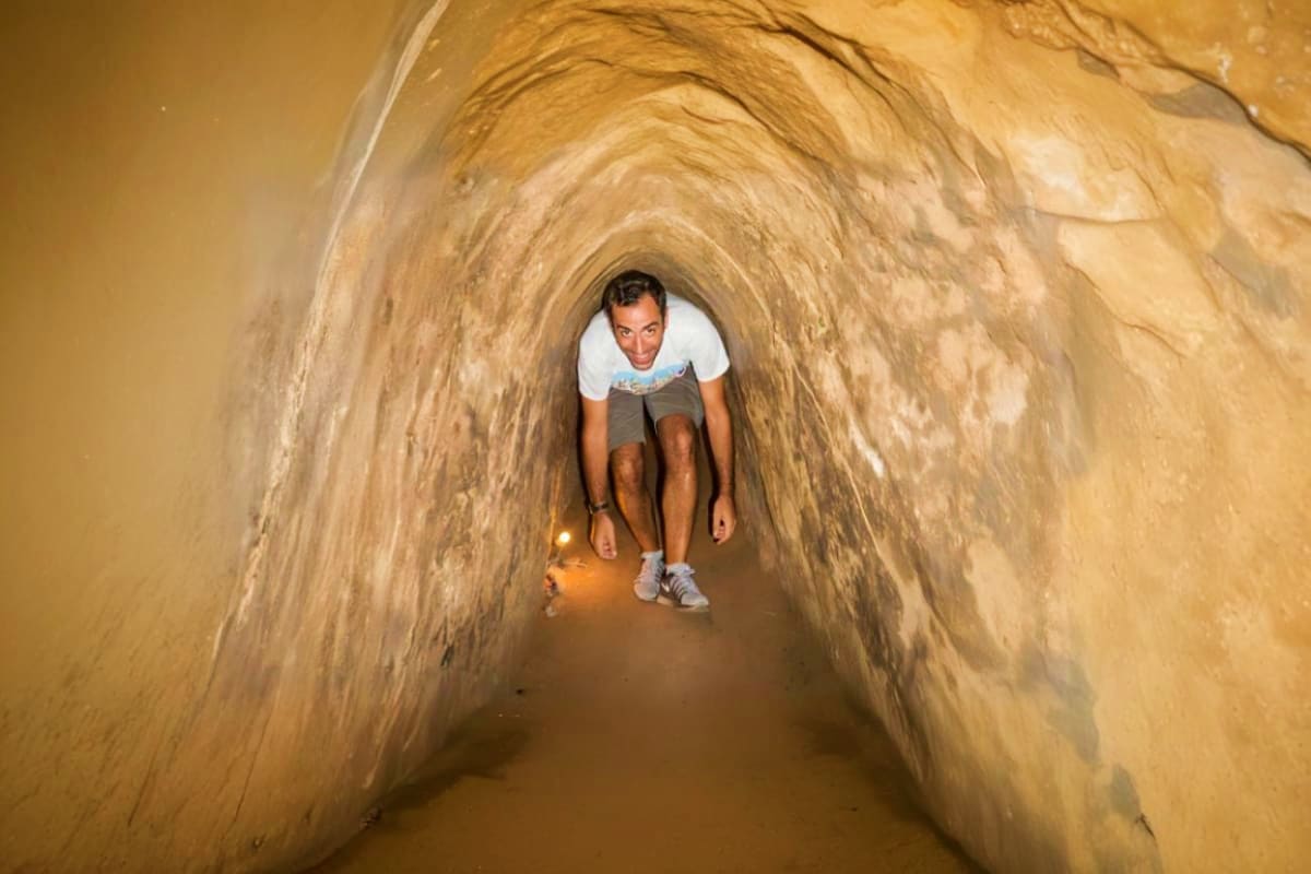 Cu Chi Tunnel Private Transfer Man crouching inside narrow underground tunnel with tan earthen walls at Cu Chi Tunnels historic site
