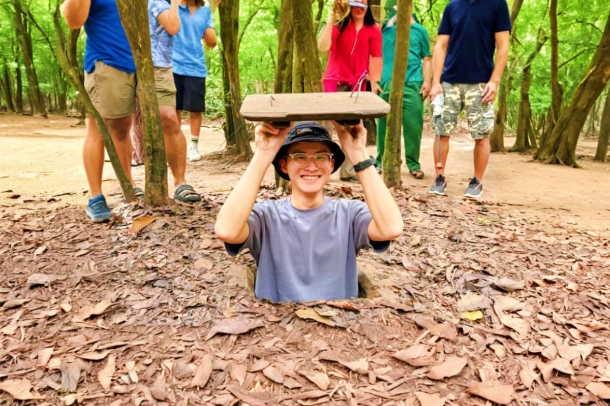 Young visitor holding wooden platform above head inside Cu Chi Tunnels with tour group standing in forest tunnel passage