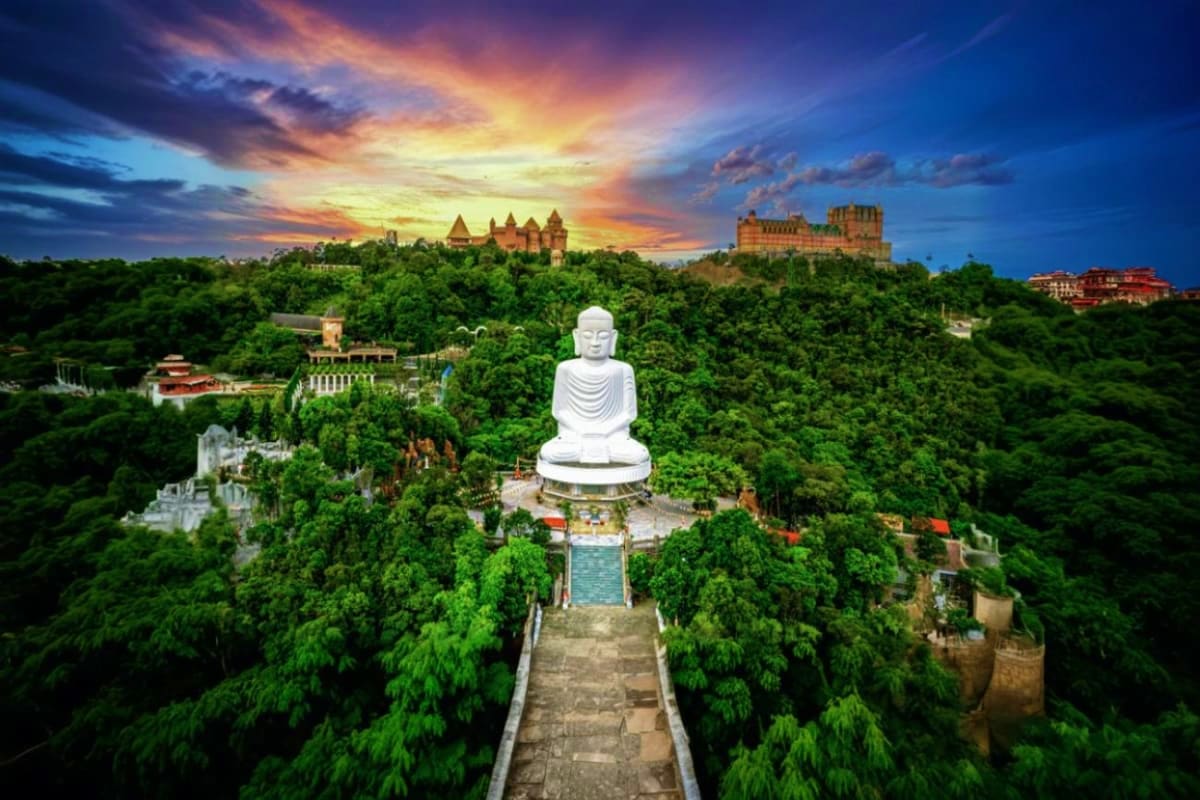 Large white Buddha statue overlooking lush green forest with castles and temples on hilltops at sunset in Da Nang, Vietnam