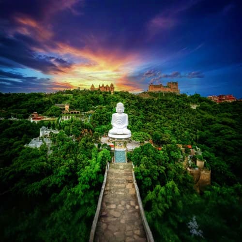 White Buddha statue on mountain pathway overlooking Danang cityscape and castle at sunset with lush green forest