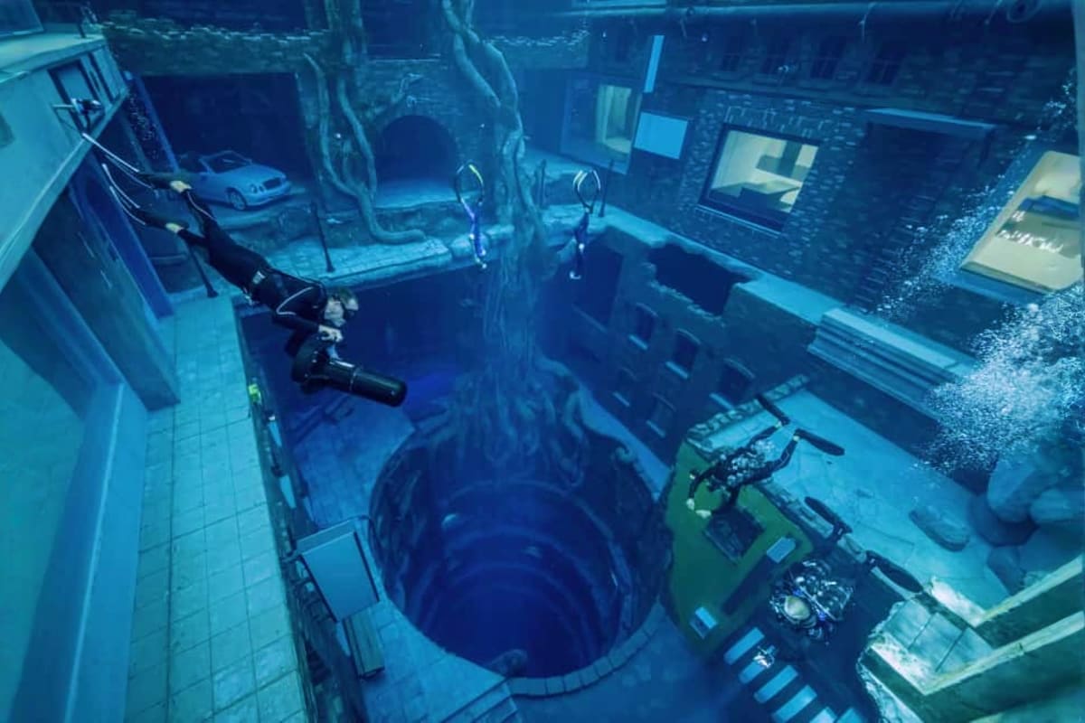 Deep Dive Dubai Underwater view of Deep Dive Dubai facility with submerged car, scuba divers, and multi-level sunken city structures in blue