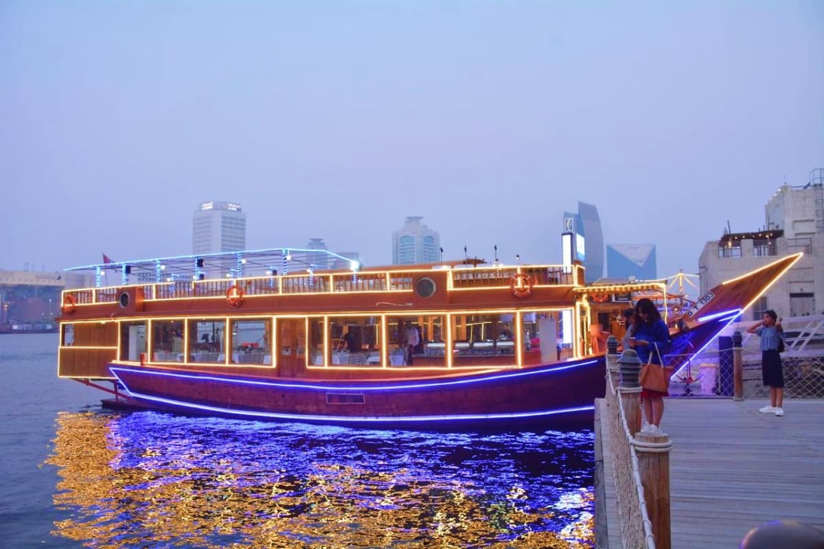 Traditional wooden dhow boat illuminated at dusk on Dubai Creek with passengers boarding, city skyline in background