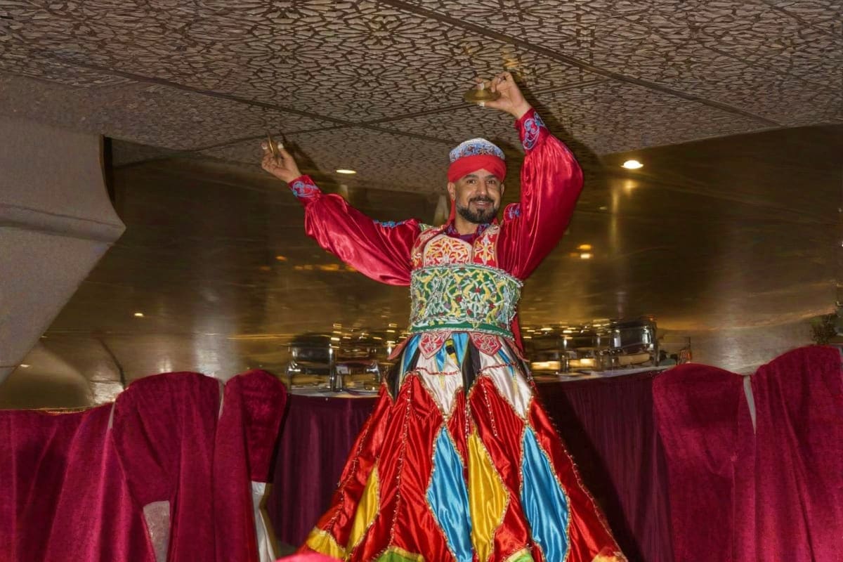 Man in traditional red and colorful Arabic clothing celebrating with arms raised on a dhow boat during creek dinner cruise
