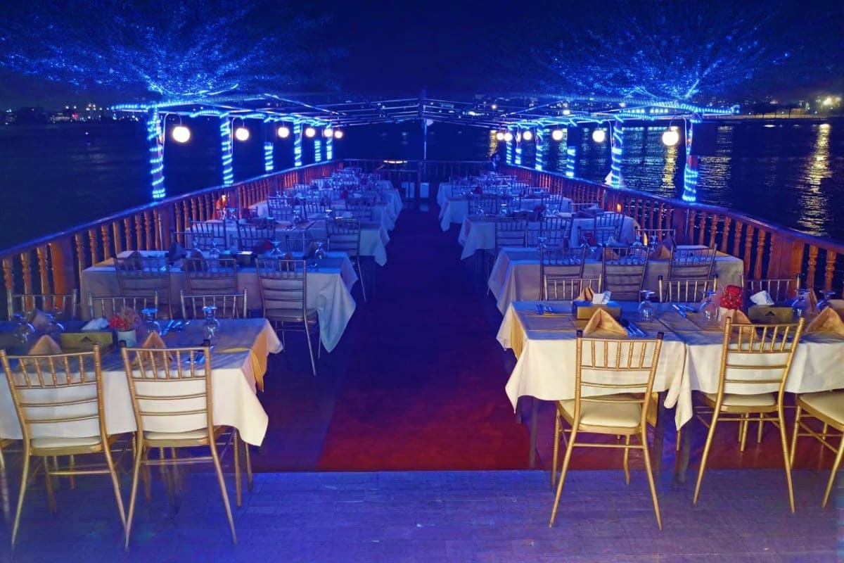 Illuminated dhow cruise dinner deck with white-clothed tables, gold chairs, and blue LED lighting along the water at night