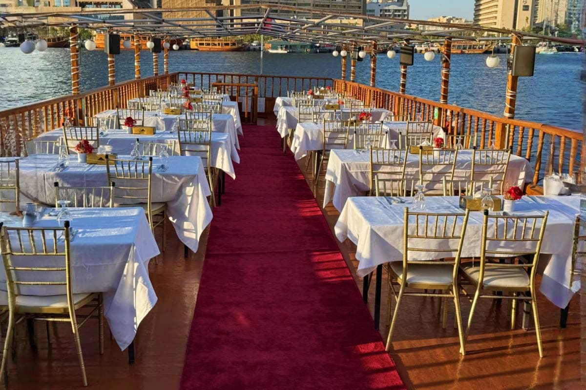 Outdoor waterfront dining setup on a dhow cruise boat with white tablecloths, gold chairs, and red carpet along the creek wit