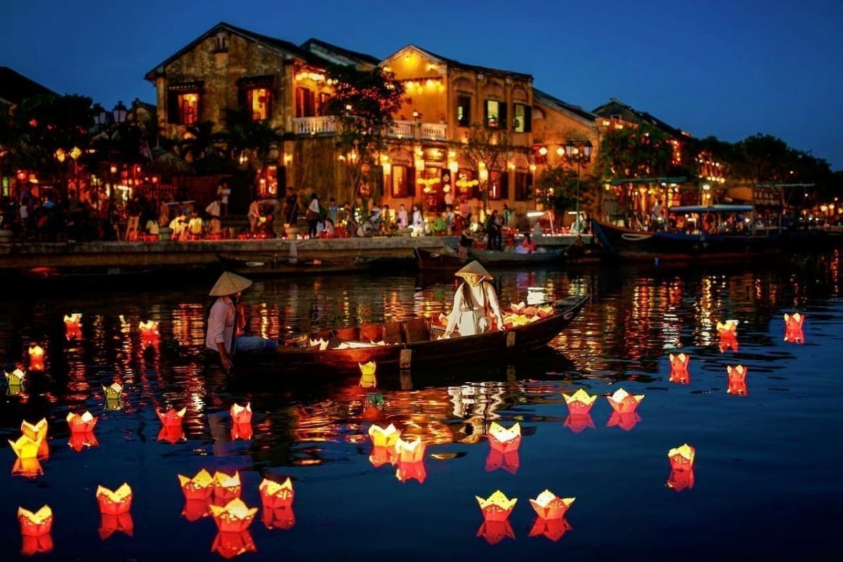 Hoi An Ancient Town waterfront at dusk with illuminated yellow buildings, traditional boats, and floating lanterns on the riv