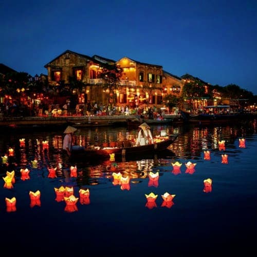 Illuminated lanterns floating on water at dusk with historic yellow buildings lit up along the waterfront in Hoi An Ancient C