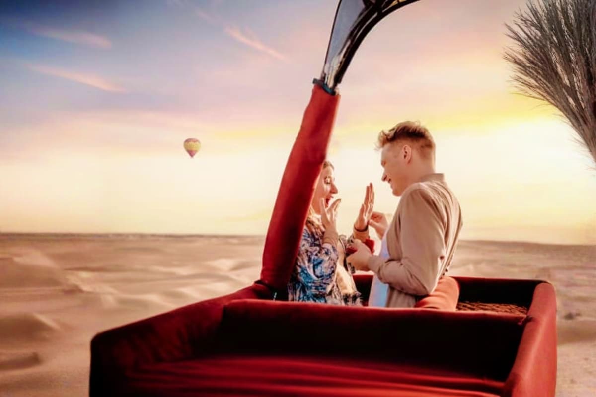 Couple in red hot air balloon basket at sunrise over Dubai desert with another balloon visible in distant sky