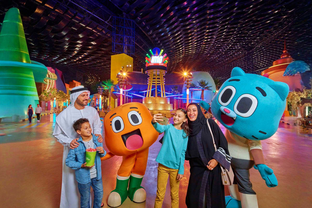 Family posing with colorful cartoon character mascots inside vibrant indoor theme park with illuminated attractions and neon 