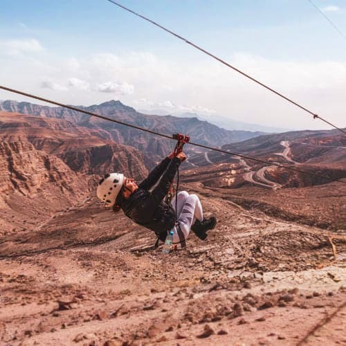 Person ziplining over desert mountains with red rock formations and winding roads in the valley below