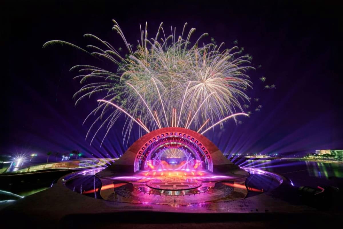 Fireworks exploding above illuminated pink and purple amphitheater stage at night by water for Kiss of the Sea Show