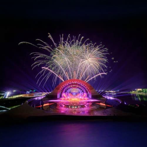 Pink illuminated amphitheater on waterfront with white fireworks bursting above night sky