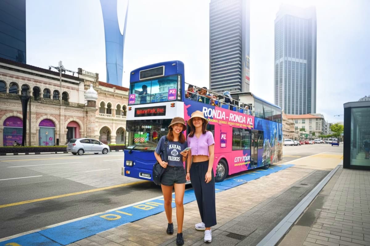 Two women posing in front of a pink and blue double-decker tour bus in Kuala Lumpur with colonial architecture and modern sky
