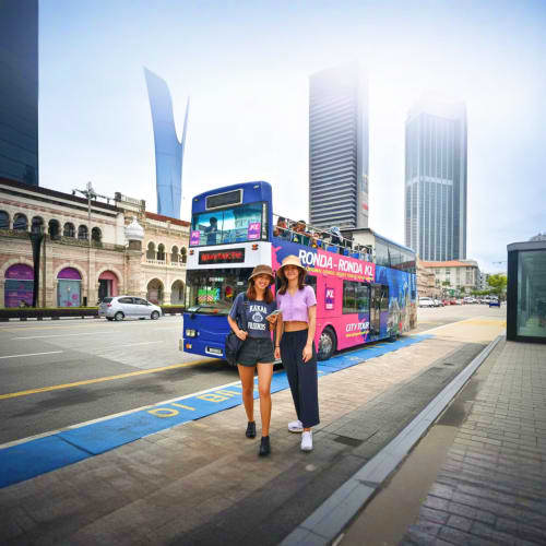 Two women walking past a colorful tour bus on a Kuala Lumpur street with modern skyscrapers and historic architecture in the 