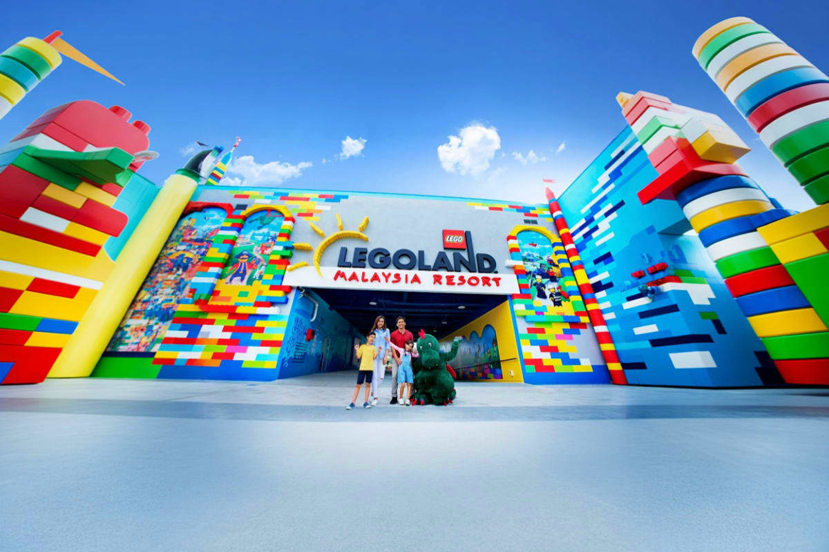 Family standing at the colorful entrance of LEGOLAND Malaysia Resort with giant LEGO brick structures and kites overhead agai