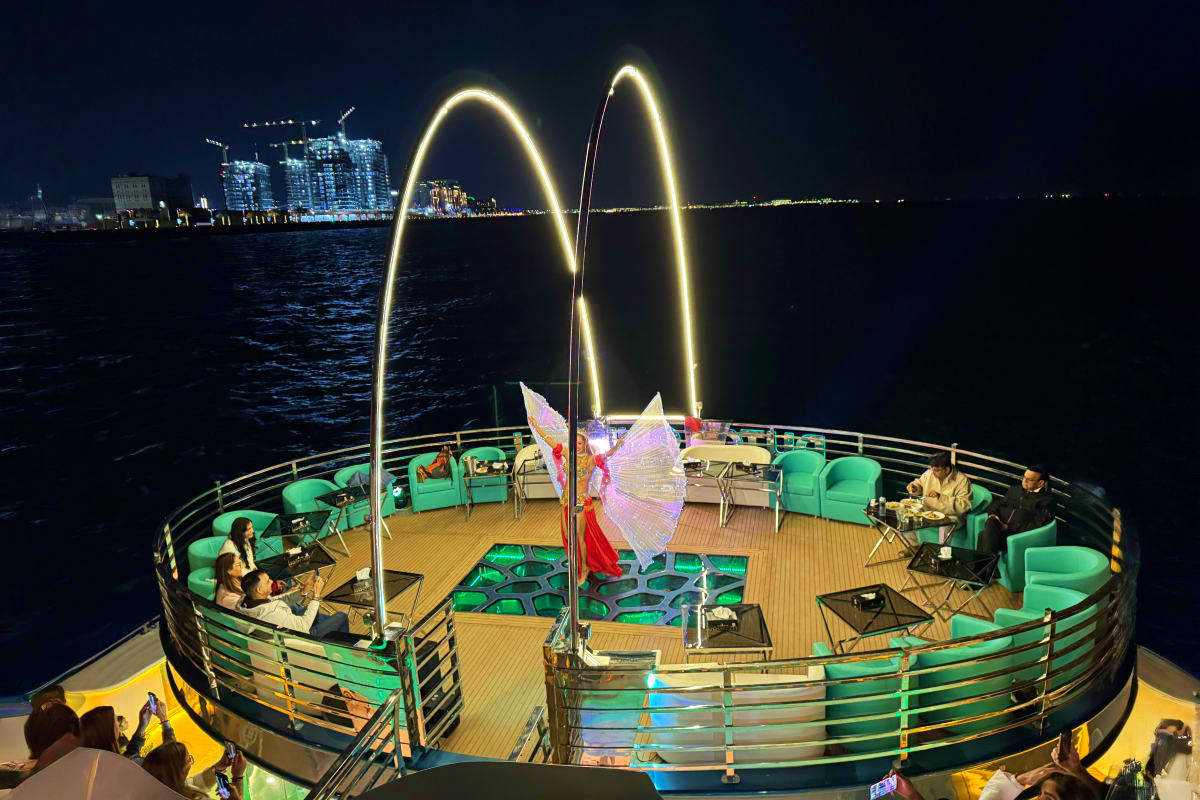 Nighttime view of Lotus Mega Yacht with illuminated arches, passengers seated in green chairs on deck, city skyline and water