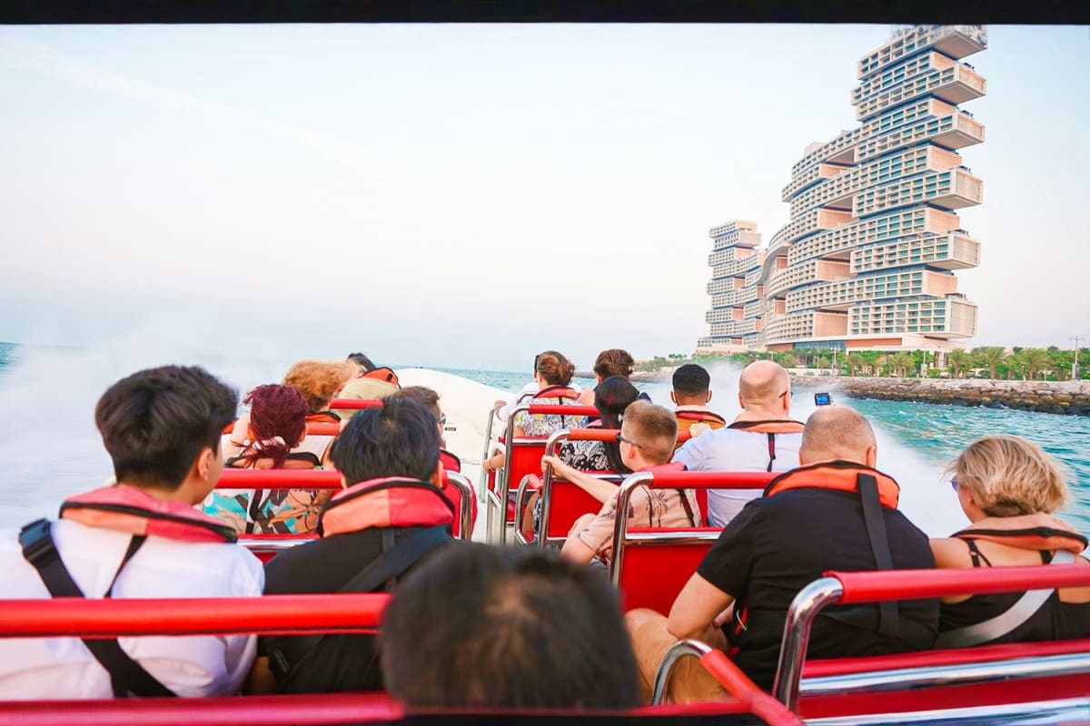 Tourists wearing life jackets seated on a red speedboat with a distinctive stepped hotel building visible in the background o