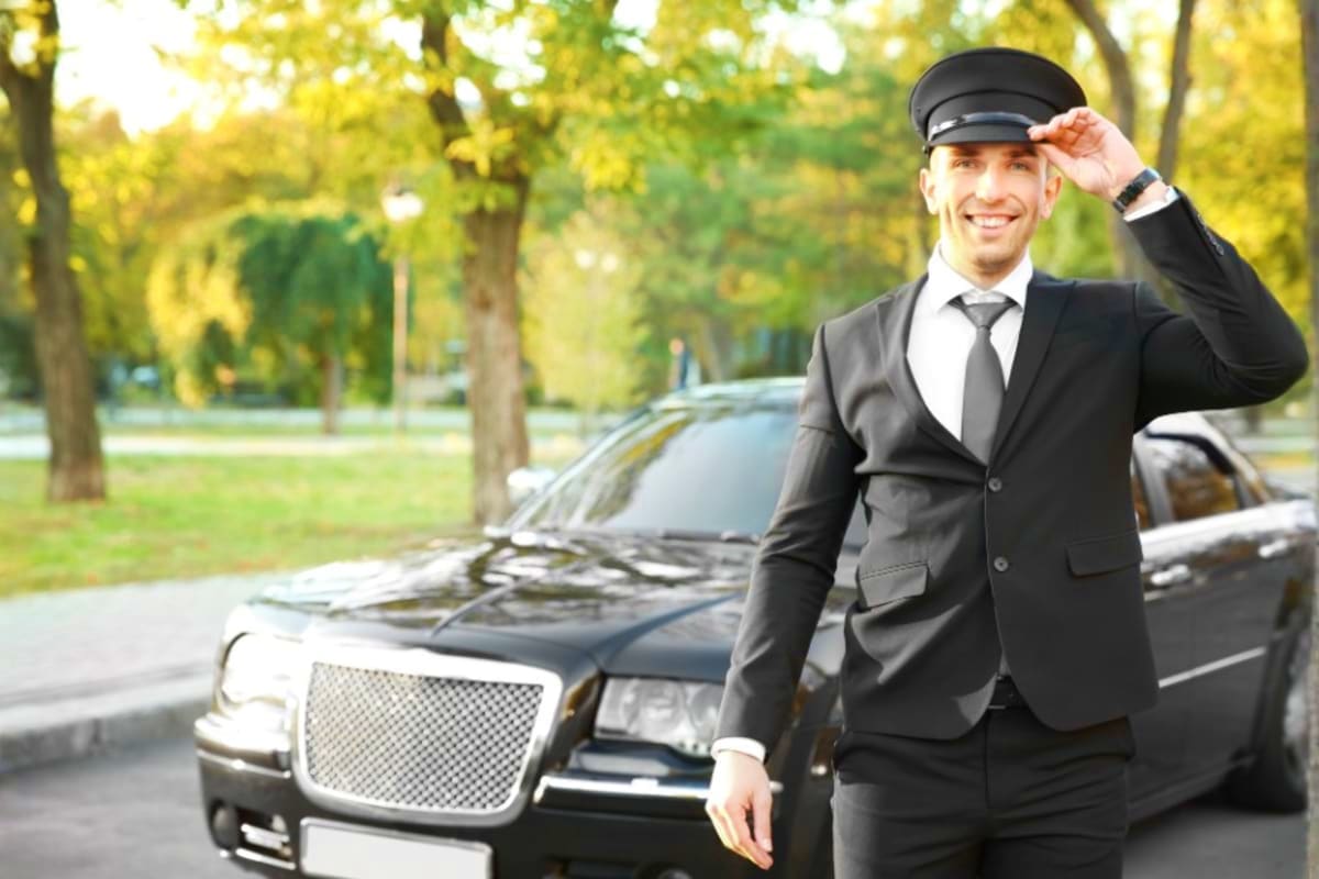 Professional chauffeur in black suit and cap standing next to luxury sedan on tree-lined street