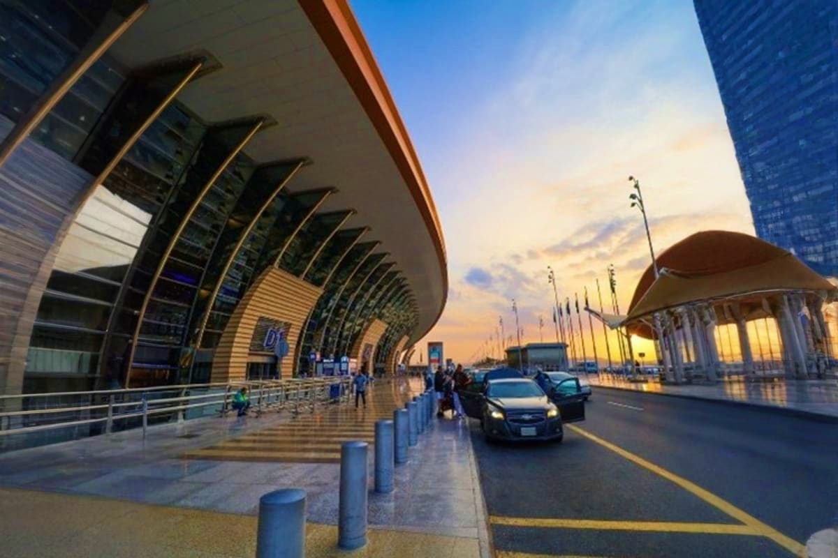 Modern airport terminal building with curved architecture at sunset, featuring passenger drop-off area and taxis