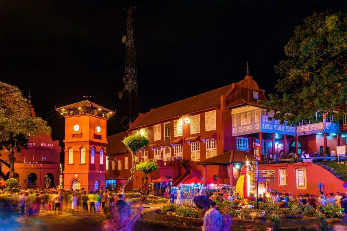 Illuminated colonial buildings in Malacca's town square at night with crowds of tourists gathering in the historic district