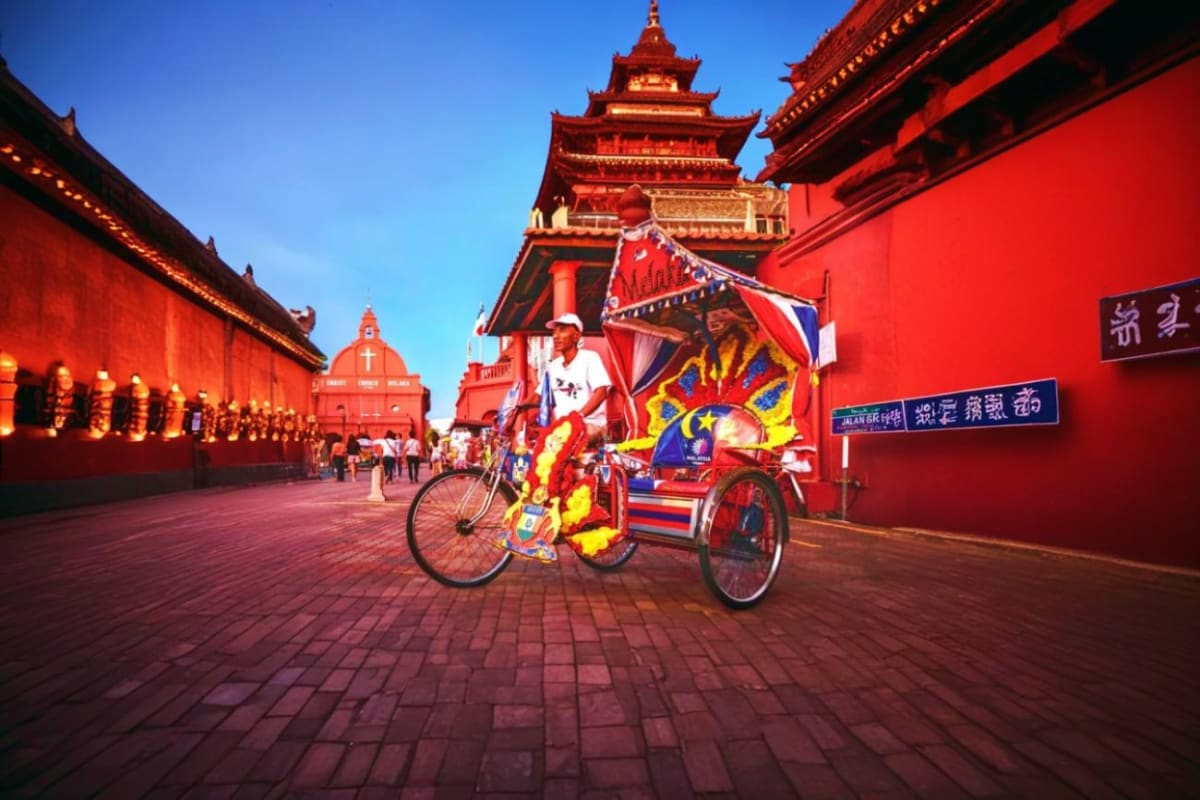 Colorful decorated trishaw with cyclist in front of red historic temple building in Malacca with blue sky