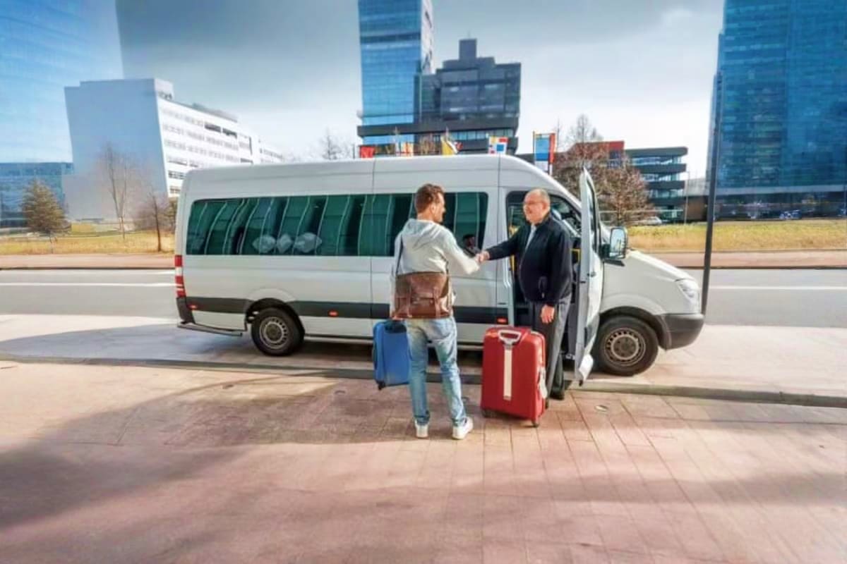 Passenger boarding a white minivan with luggage at an urban transit station with modern office buildings in background