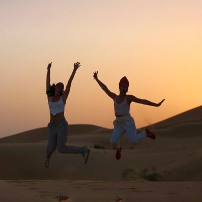 Morning Safari Two women jumping joyfully on desert sand at sunset during a morning desert safari tour