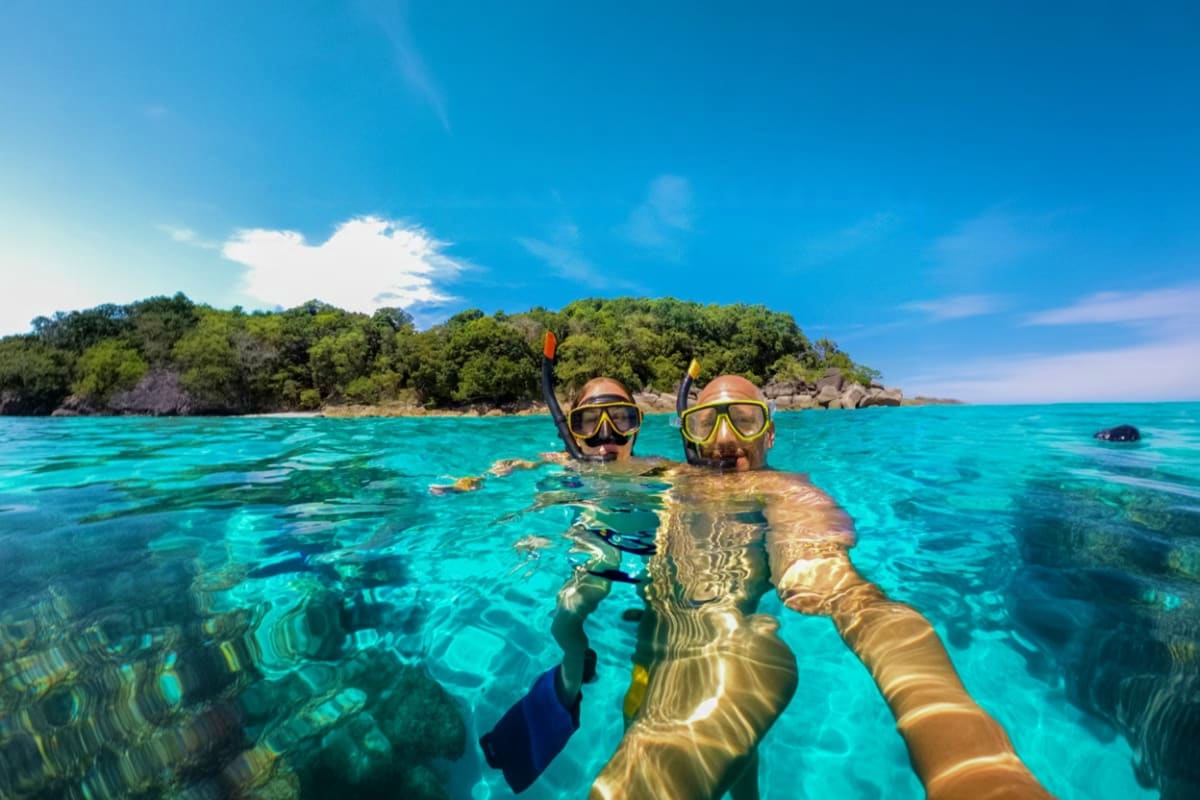 Nusa Penida Day Tour And Snorkeling Depart from Nusa Penida Two snorkelers in wetsuits taking a selfie in crystal clear turquoise water with tropical island and vegetation in background