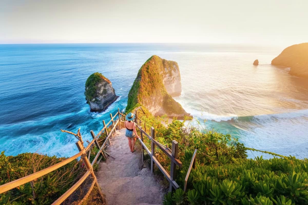 Hiker on wooden boardwalk overlooking Kelingking Beach cliff rock formations and turquoise ocean at sunset, Nusa Penida Bali