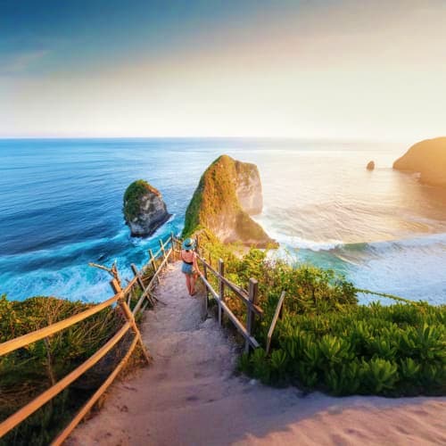 Wooden railing pathway overlooking Kelingking Beach with dramatic rock formations and turquoise ocean waters at sunset, Nusa 