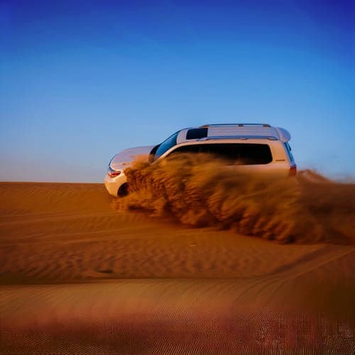 White SUV driving through desert sand dunes creating dust cloud under clear blue sky