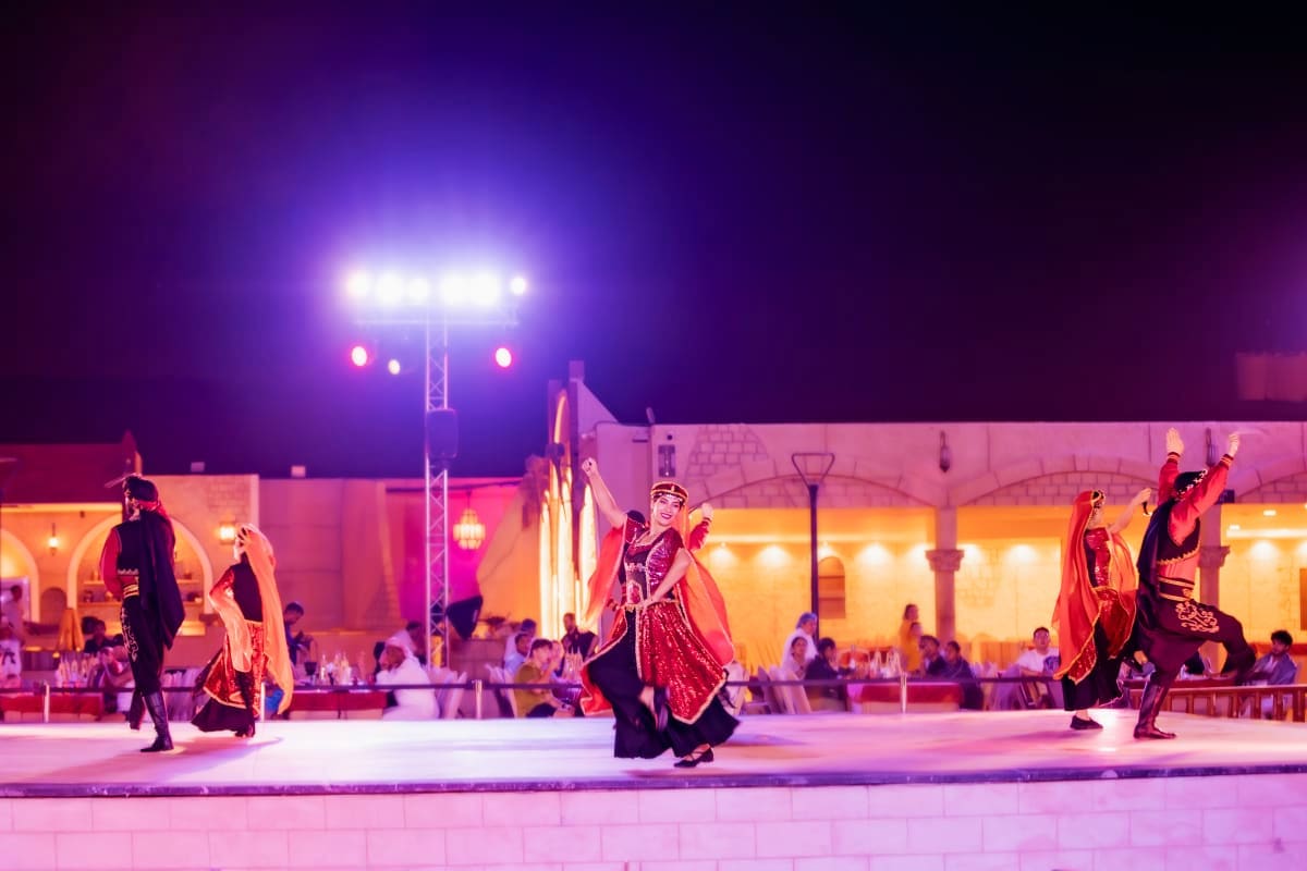 Traditional Middle Eastern performers in red and gold costumes dancing on illuminated stage at night venue with audience seat