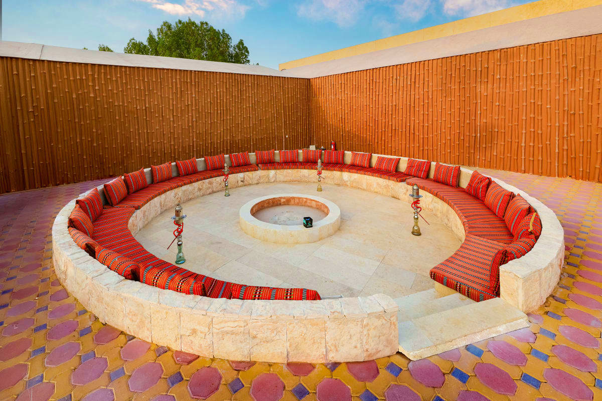 Circular stone seating area with red patterned cushions and central fire pit on tiled courtyard, surrounded by brick walls un