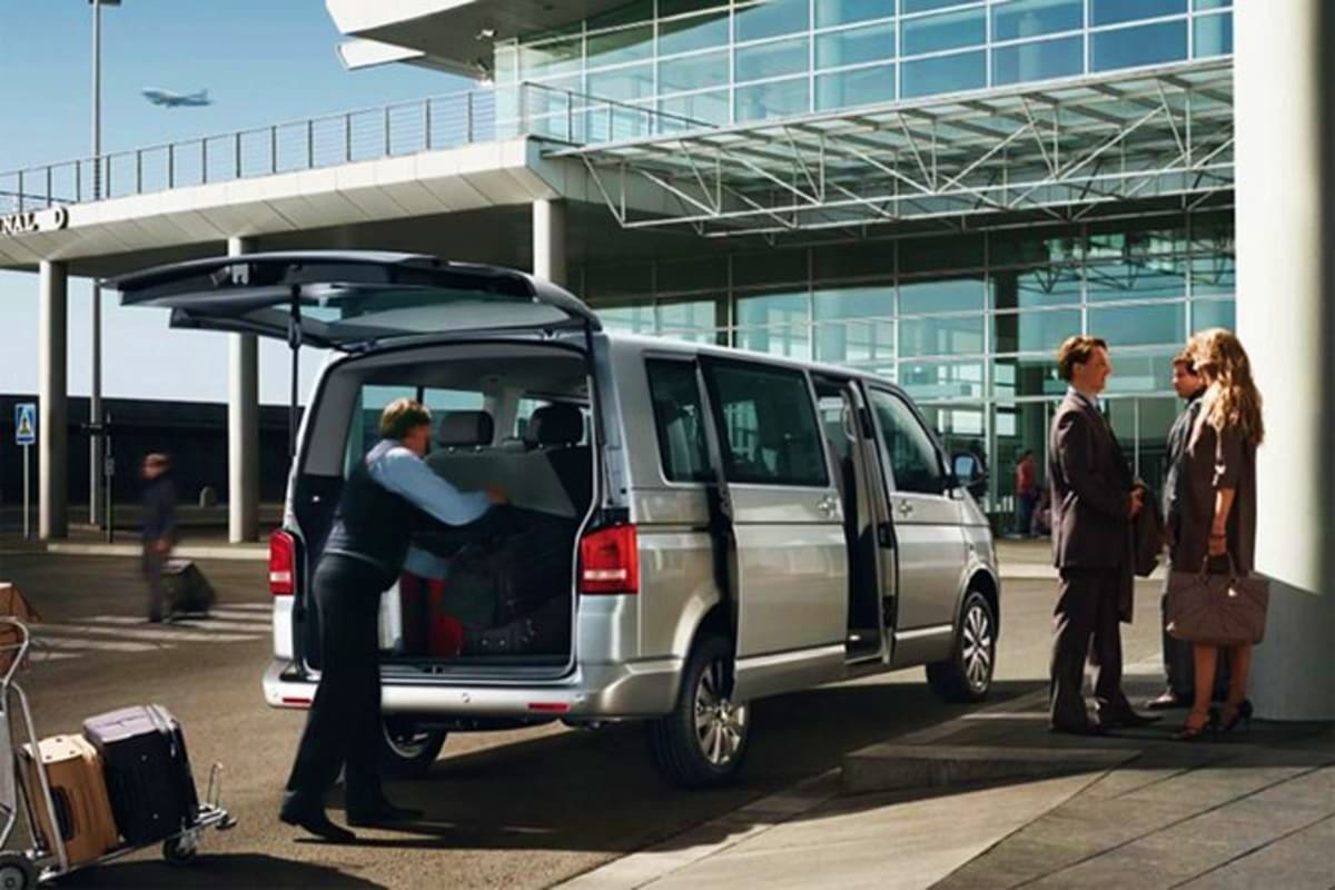 Business travelers with luggage boarding silver minivan at modern airport terminal with departing airplane visible