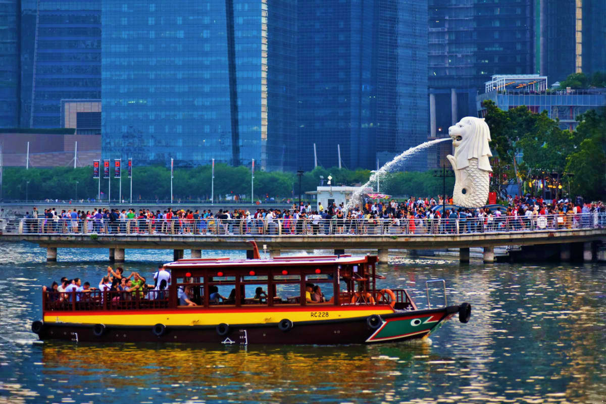 River Cruise Singapore Tourist boat with red and yellow stripe cruising past Merlion statue and modern skyscrapers in Singapore River